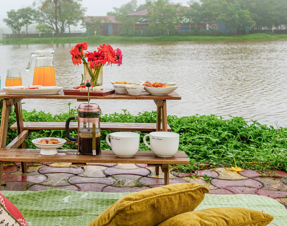 Lakeside homestay setup in Lonavala with coffee, snacks, and flowers on a wooden bench by the water