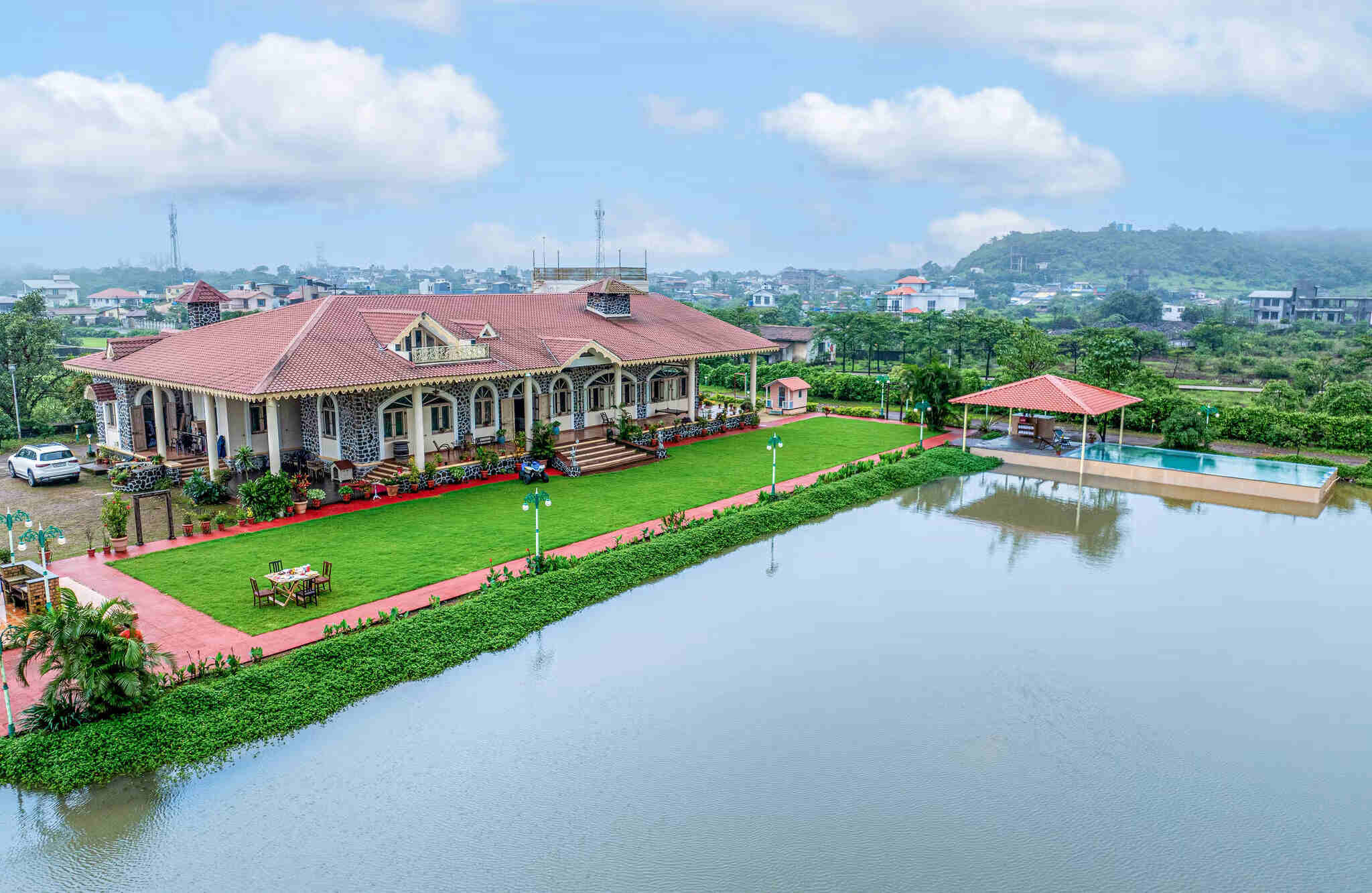 Luxurious villa in Lonavala with a private swimming pool, large lawn, and lakeside view under a cloudy sky
